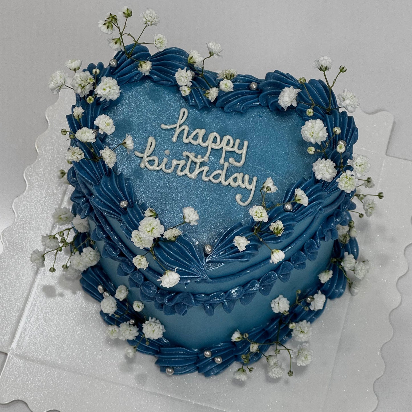Pastel azul con acabado brillante y flores blancas, decoración elegante en crema con texto “Happy Birthday”