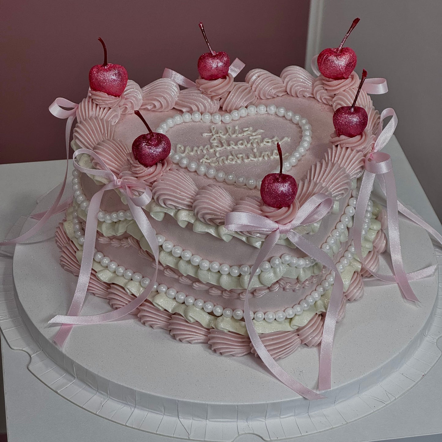 Pastel rosa con forma de corazón, cerezas con glitter comestible y lazos, ideal para celebraciones elegantes.