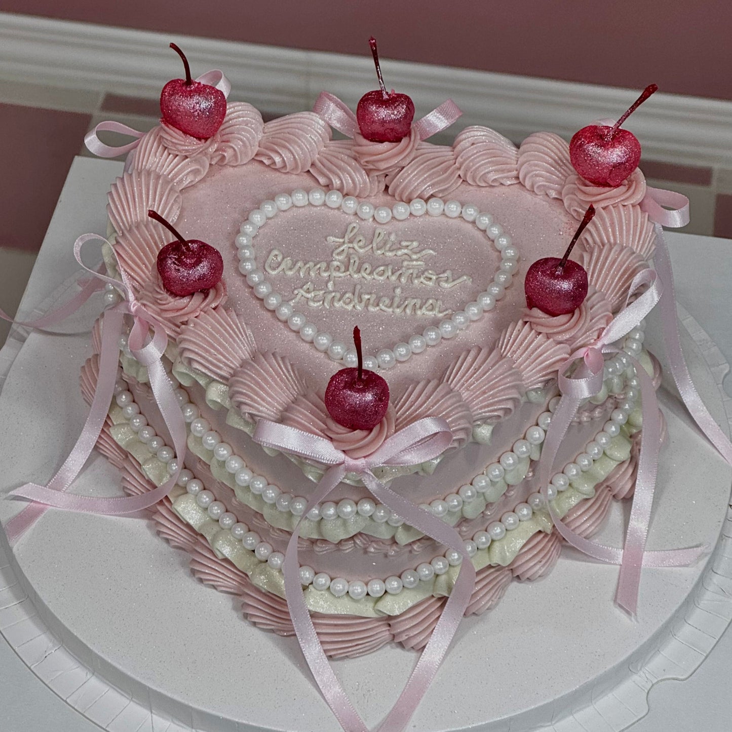 Tarta en forma de corazón decorada con cerezas brillantes, crema rosa, perlas y mensaje “Feliz cumpleaños”