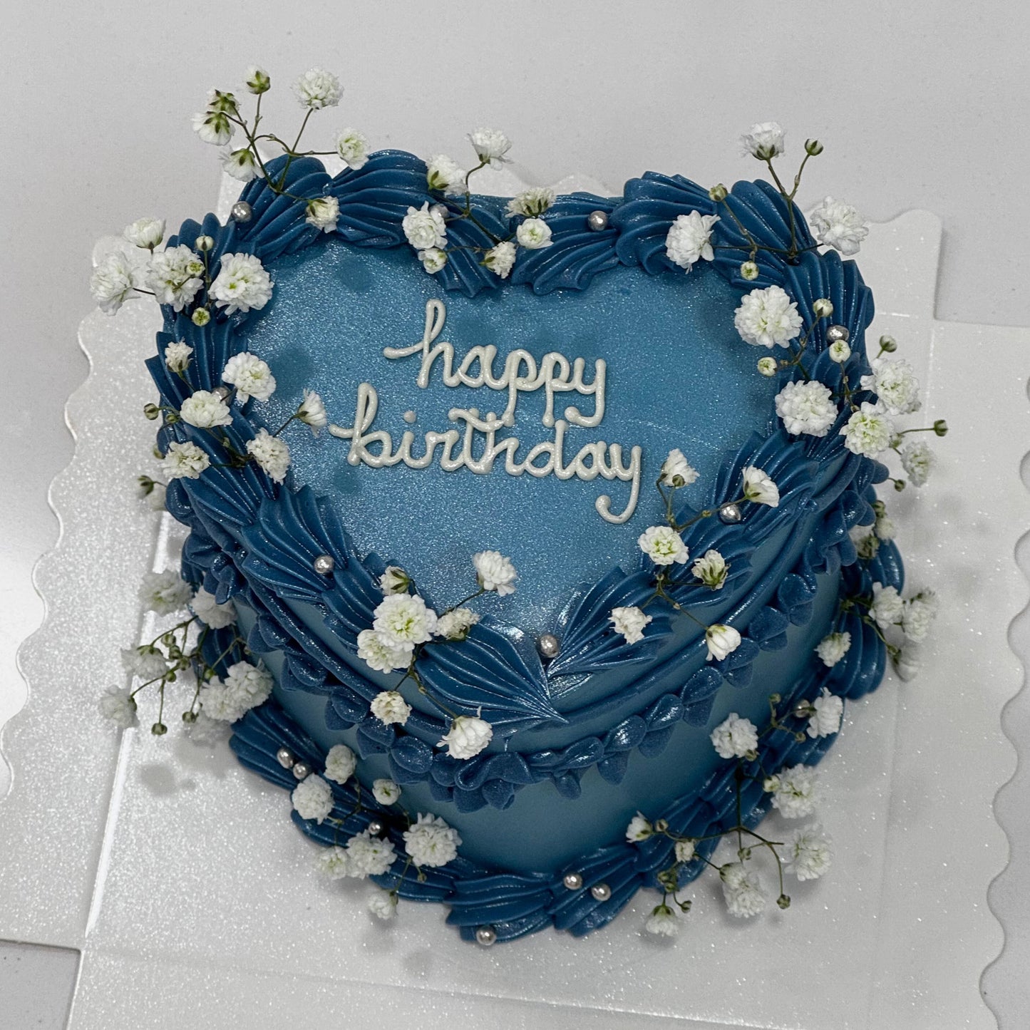 Tarta azul en forma de corazón con mensaje “Happy Birthday”, decorada con crema ondulada y flores blancas naturales.