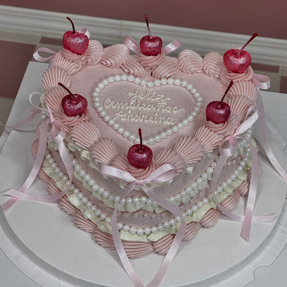 Tarta en forma de corazón decorada con cerezas brillantes, crema rosa, perlas y mensaje “Feliz cumpleaños”